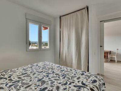 Chambre lumineuse avec lit à motifs, fenêtre offrant des vues urbaines et une armoire discrète couverte par un rideau clair, avec accès à une pièce adjacente.