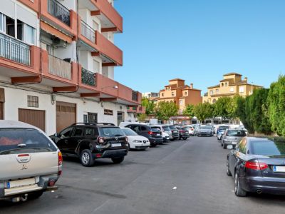 Espace de stationnement extérieur avec plusieurs véhicules, bordé par un immeuble résidentiel aux balcons orange et des structures ocre traditionnelles en arrière-plan sous un ciel clair.