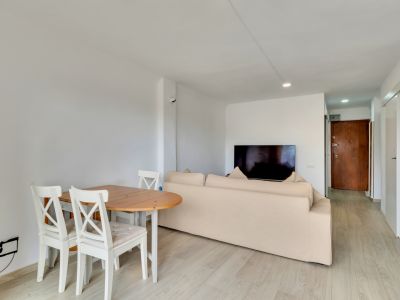 Espace salon-salle à manger ouvert avec table en bois clair, chaises blanches et canapé beige. Télévision murale et sol stratifié clair menant à une porte d'entrée en bois foncé.