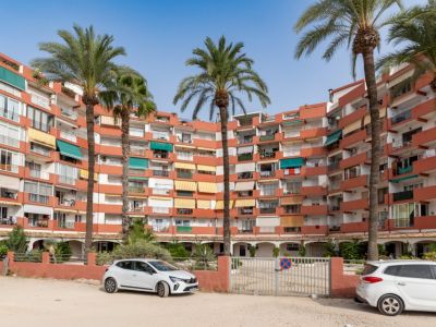 Immeuble d'appartements incurvé avec façade terre cuite et balcons aux stores variés. De grands palmiers en premier plan, avec un parking extérieur et un portail.