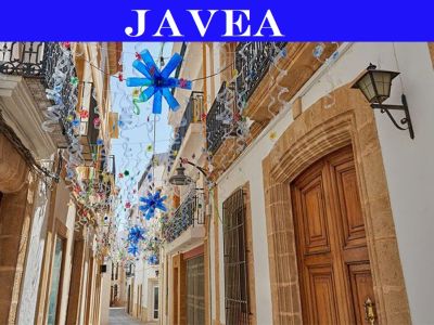 Rue étroite du centre historique de Javea, avec des portes en bois encadrées de pierre, des balcons en fer forgé et des décorations festives aériennes en forme d'étoiles bleues.