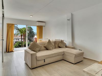 Salon lumineux avec canapé d'angle beige, sol effet bois clair et accès à un balcon offrant des vues sur les palmiers et des rideaux jaunes.