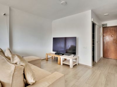 Séjour lumineux avec canapé sectionnel beige et TV sur des tables en bois clair. Le sol stratifié clair mène à la porte d'entrée en bois foncé et à un espace de rangement étagère.