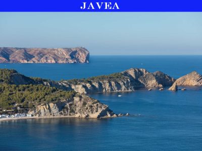 Vue panoramique de la côte rocheuse de Jávea, avec des falaises boisées de pins, une crique isolée aux maisons blanches et des îlots rocheux dans la Méditerranée bleue, un phare au loin.