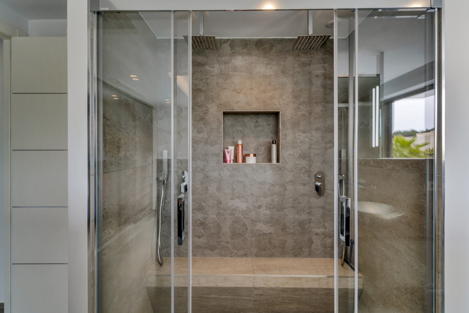 Cabine de douche moderne avec parois vitrées, carrelage gris effet pierre et deux pommes de douche pluie. Comprend un banc intégré, une niche encastrée et des rangements blancs. Vue partielle sur l'extérieur.