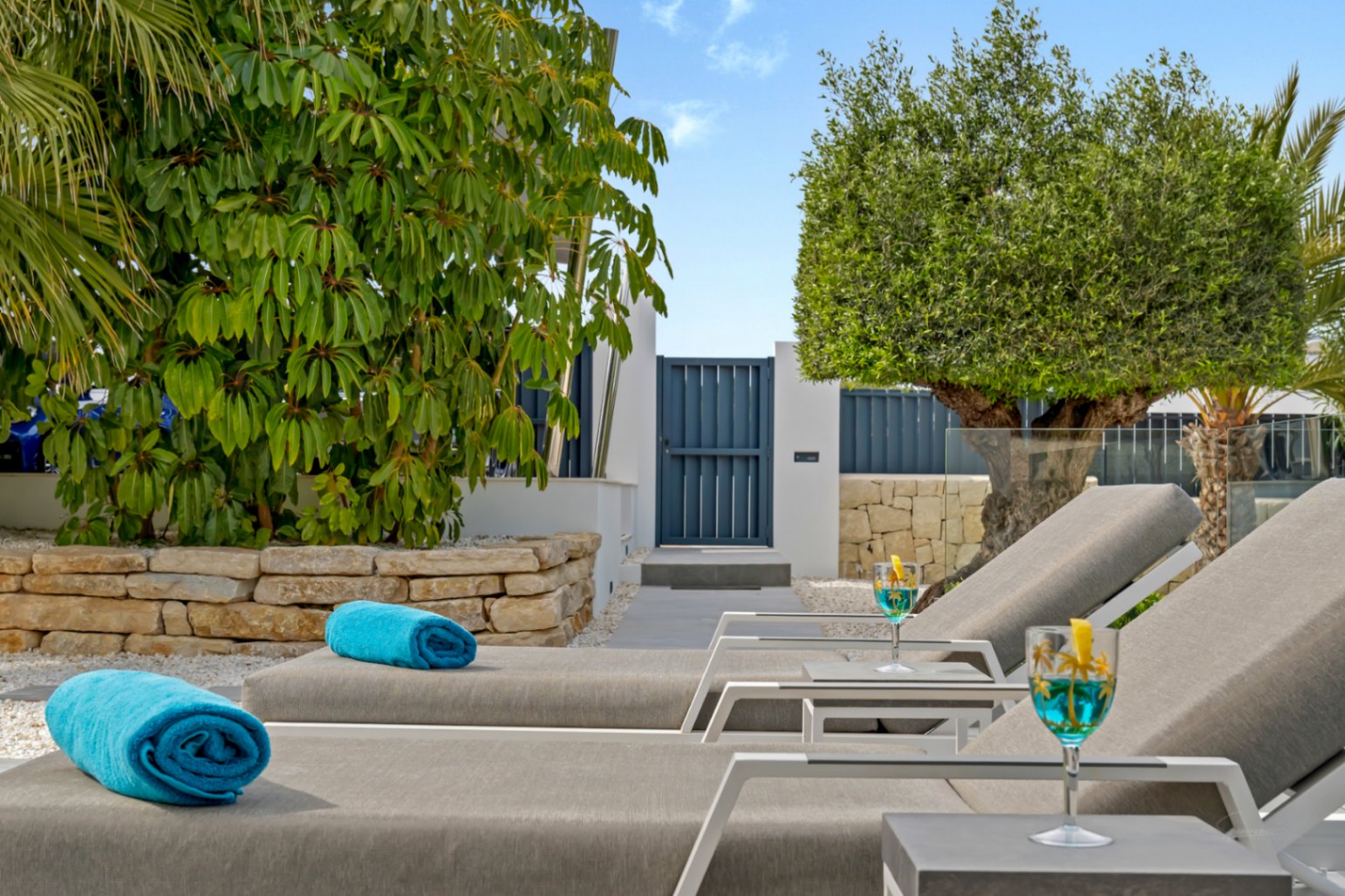 Chaises longues modernes avec serviettes bleues et cocktails rafraîchissants, dans un jardin méditerranéen luxuriant avec mur en pierre et portail bleu élégant.