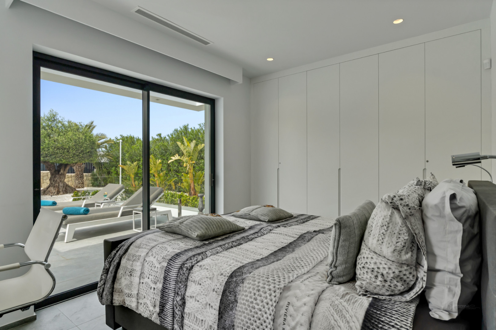 Chambre moderne avec accès direct à une terrasse privée. De grandes baies vitrées révèlent des chaises longues et une végétation méditerranéenne luxuriante, avec des armoires intégrées.