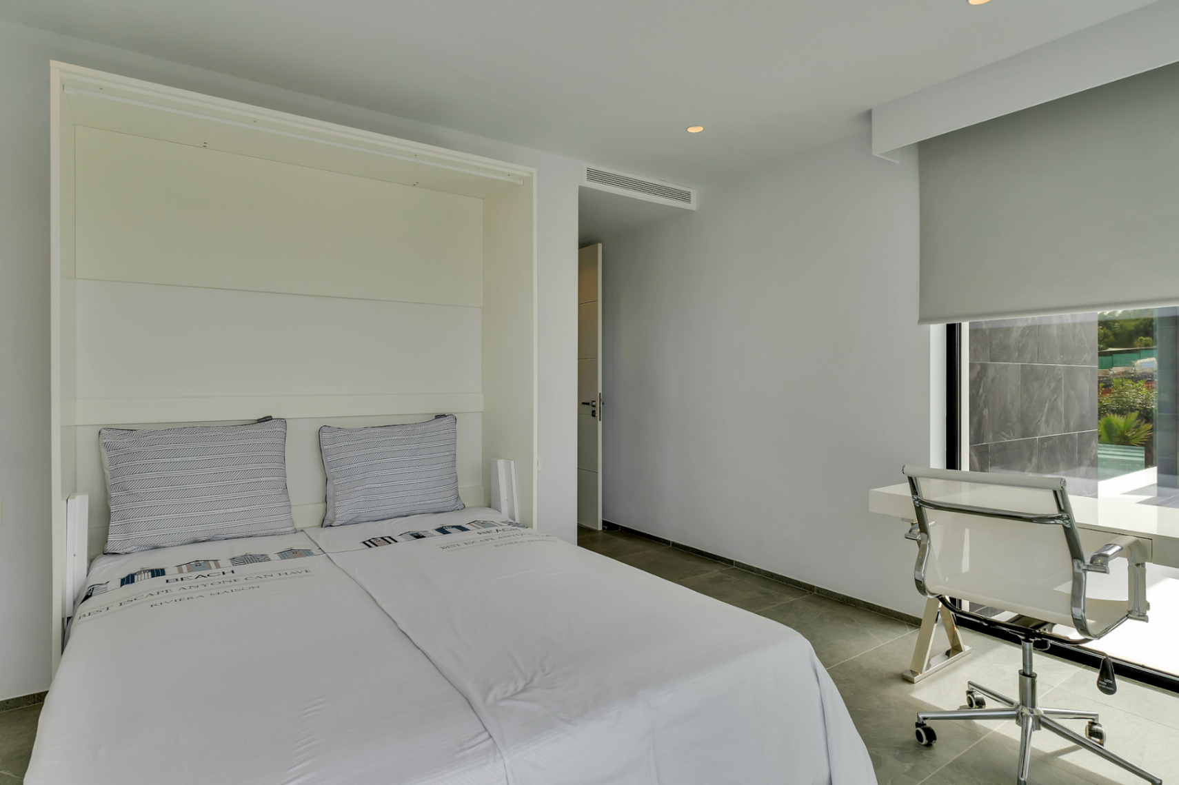 Chambre moderne avec lit encastré et espace bureau près d'une fenêtre, offrant une vue partielle sur une terrasse carrelée extérieure et de la verdure. Sol en carrelage gris.