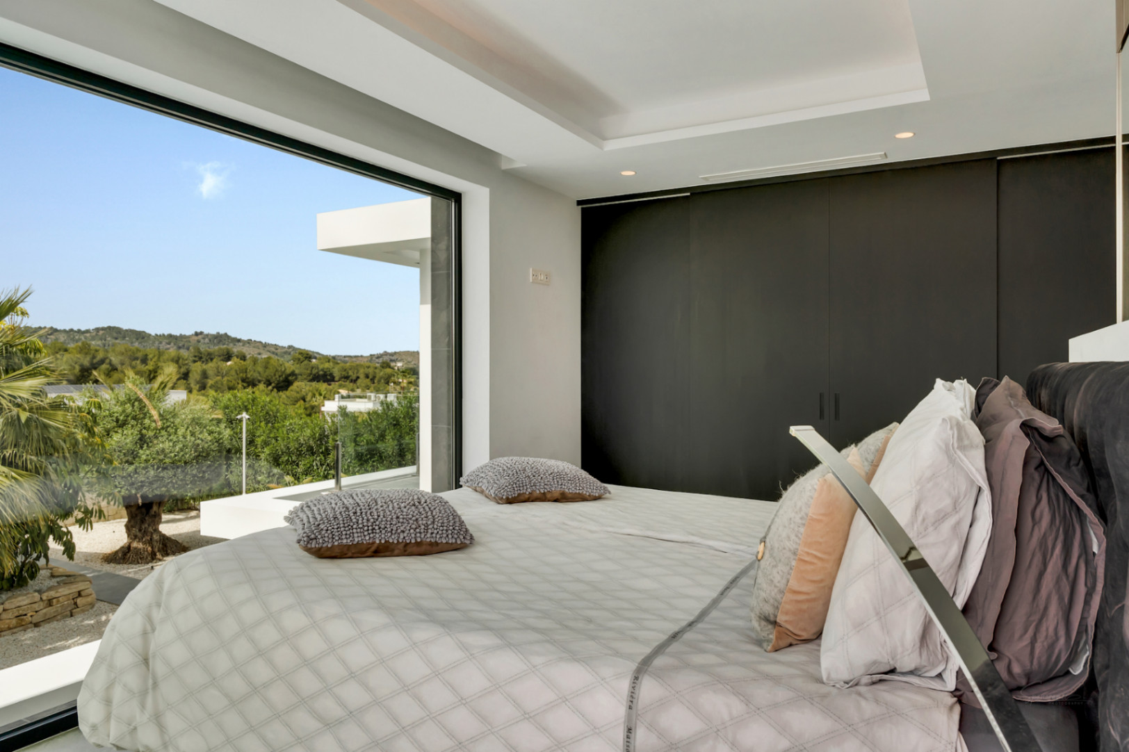 Chambre moderne avec une fenêtre du sol au plafond offrant une vue panoramique sur les collines verdoyantes. Des armoires encastrées sombres et un éclairage discret complètent l'espace.