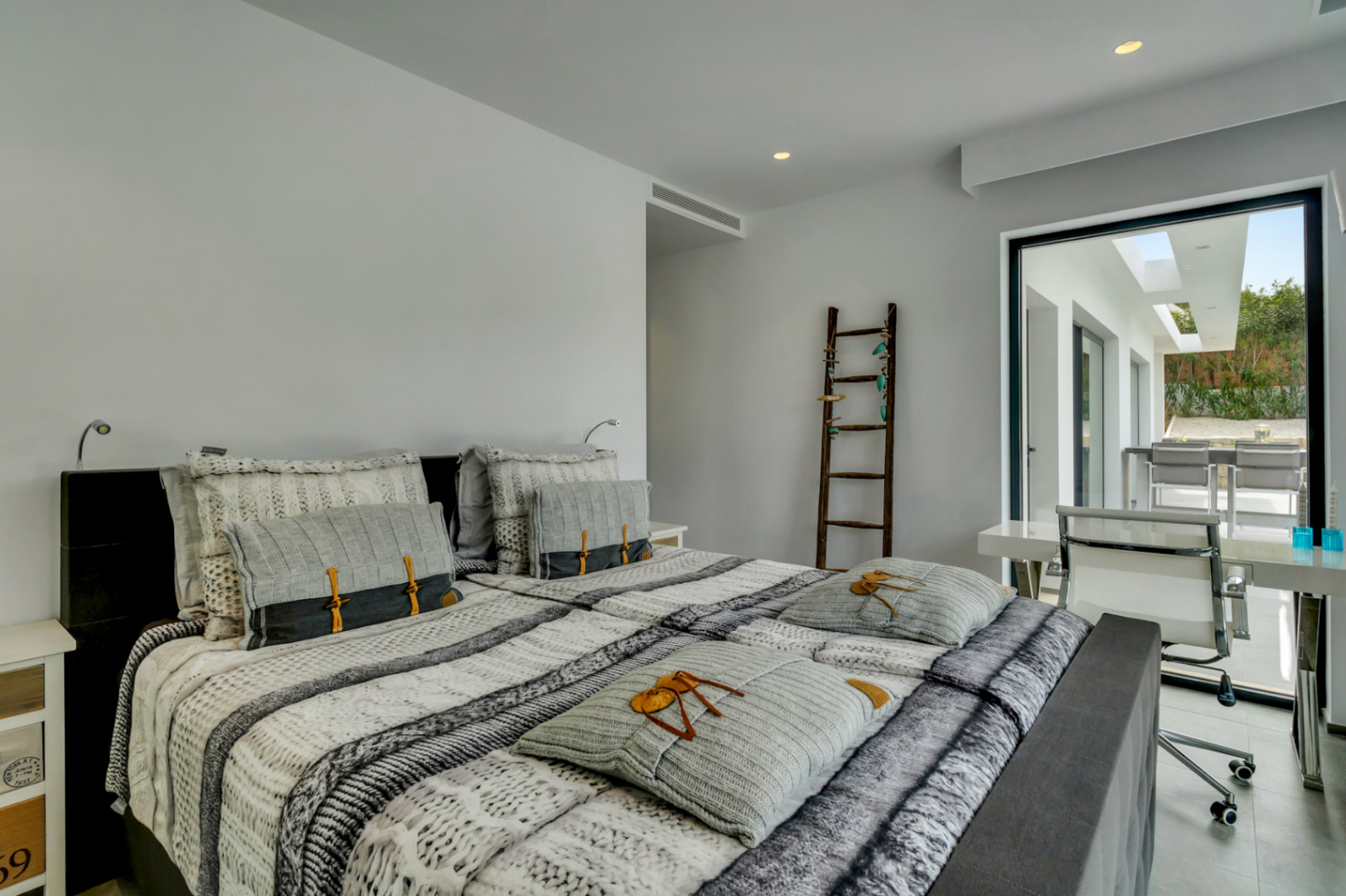 Chambre principale moderne avec lit double, espace bureau intégré et accès direct à une terrasse extérieure, éclairée par la lumière naturelle et des spots encastrés.