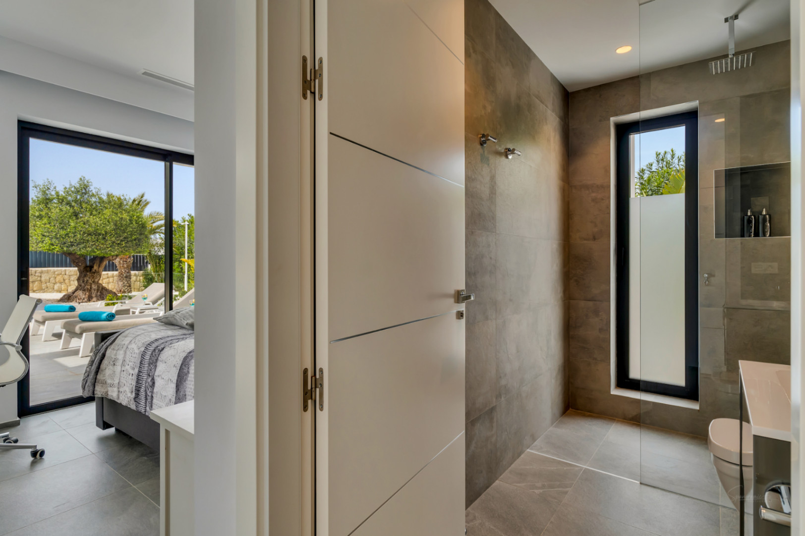 Entrée d'une salle de bain moderne avec douche à l'italienne carrelée et fenêtre dépolie, jouxtant une chambre offrant un accès direct à une terrasse arborée.