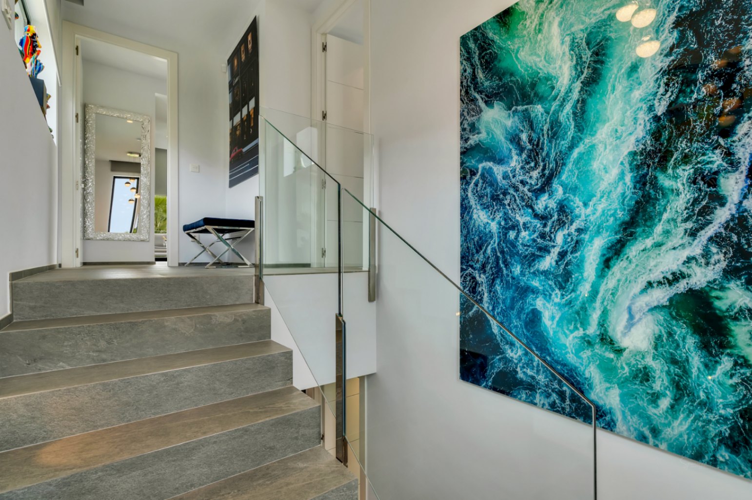Escalier intérieur en pierre grise avec rampe en verre, rehaussé par une grande œuvre d'art abstraite bleu-vert et un couloir avec miroir décoratif.