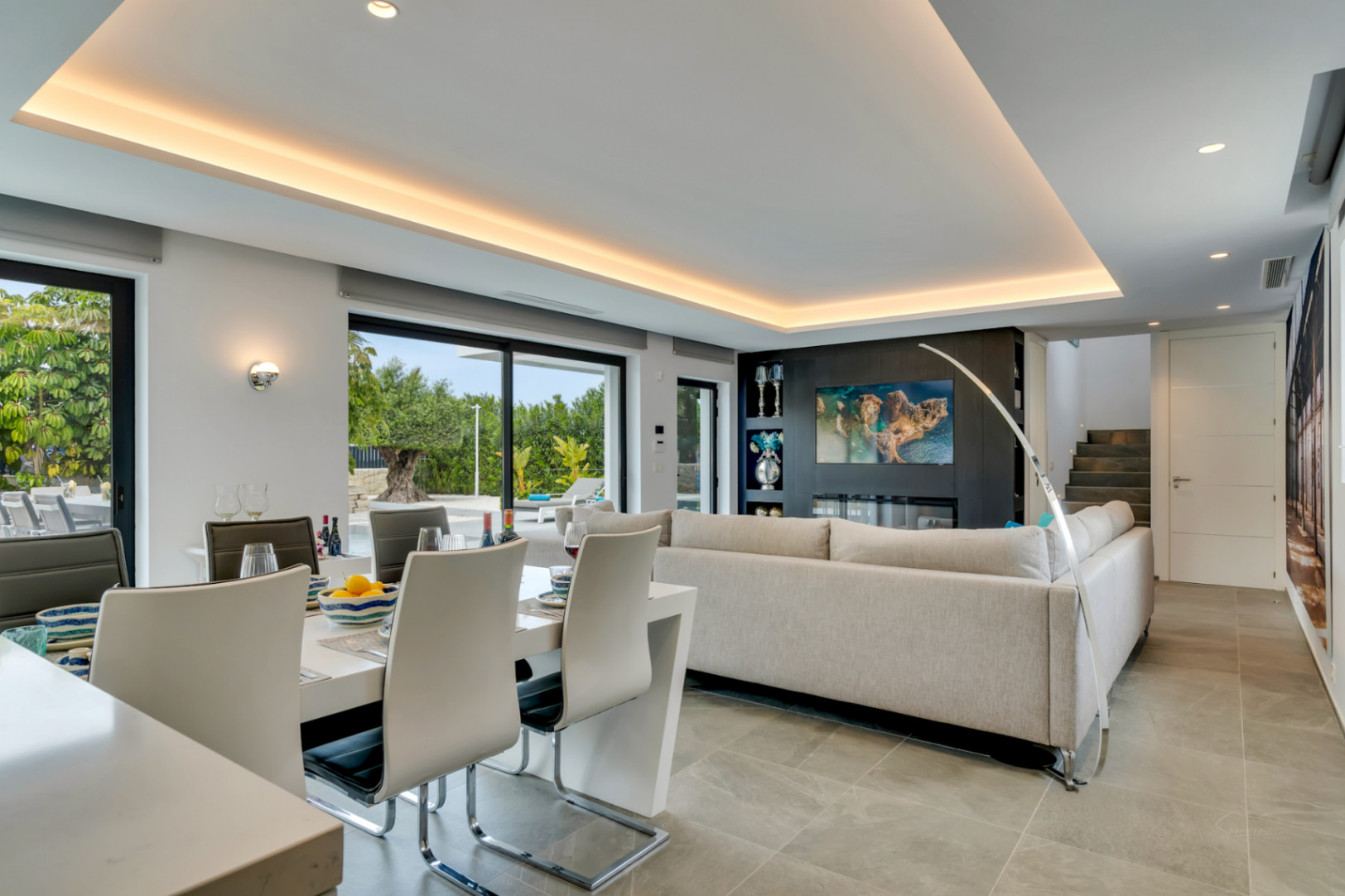 Espace de vie et salle à manger ouvert moderne avec table blanche élégante, canapé gris clair et mur média intégrant une cheminée électrique. Larges baies vitrées donnent sur le jardin.