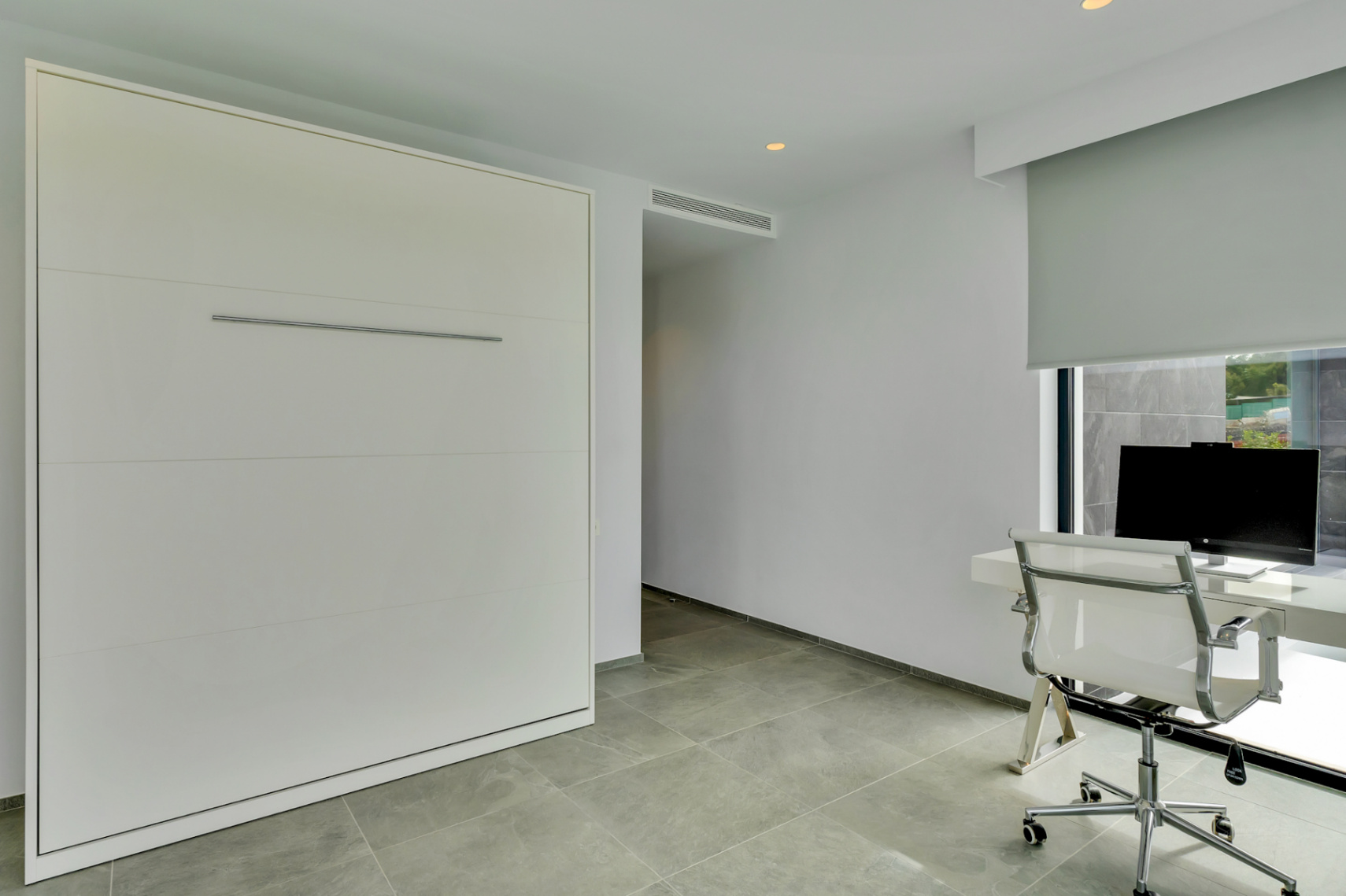 Espace multifonctionnel avec lit escamotable intégré, bureau moderne devant une fenêtre avec store et carrelage gris au sol.