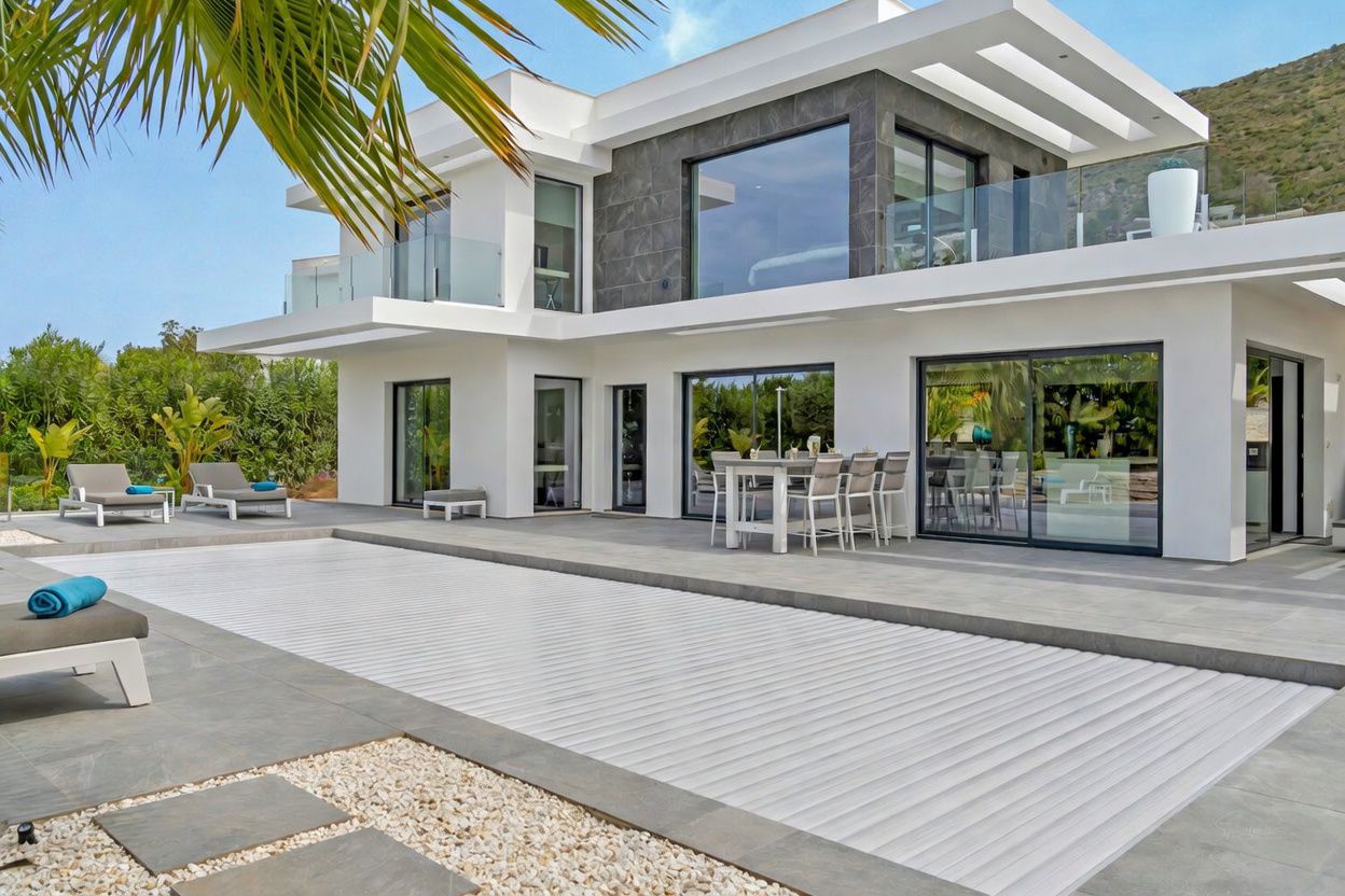 Extérieur de villa moderne avec piscine couverte à lames blanches et vaste terrasse carrelée grise. Comprend un espace repas extérieur et de grandes baies vitrées reflétant l'environnement.