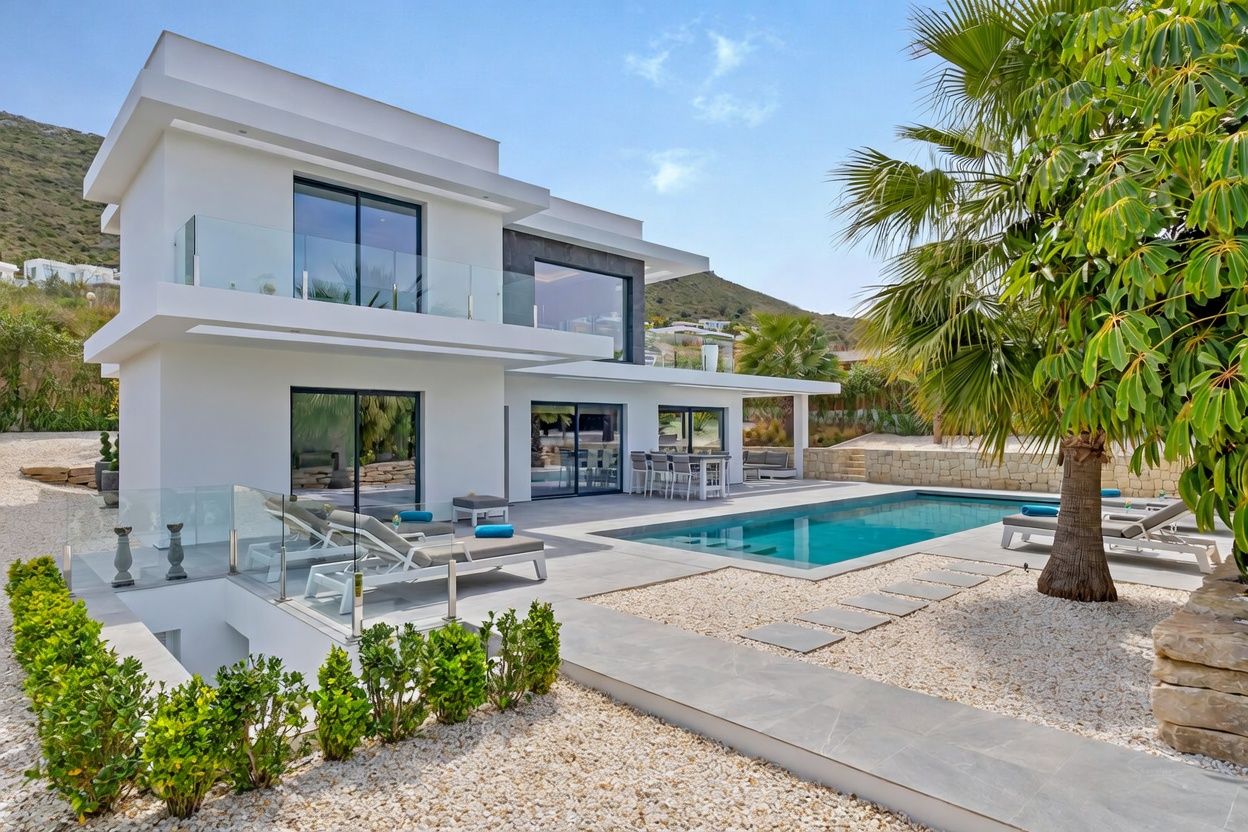 Extérieur d'une villa blanche moderne avec piscine rectangulaire, terrasse carrelée grise et salle à manger extérieure couverte. Aménagement paysager de palmiers et gravier blanc, vue sur la montagne.