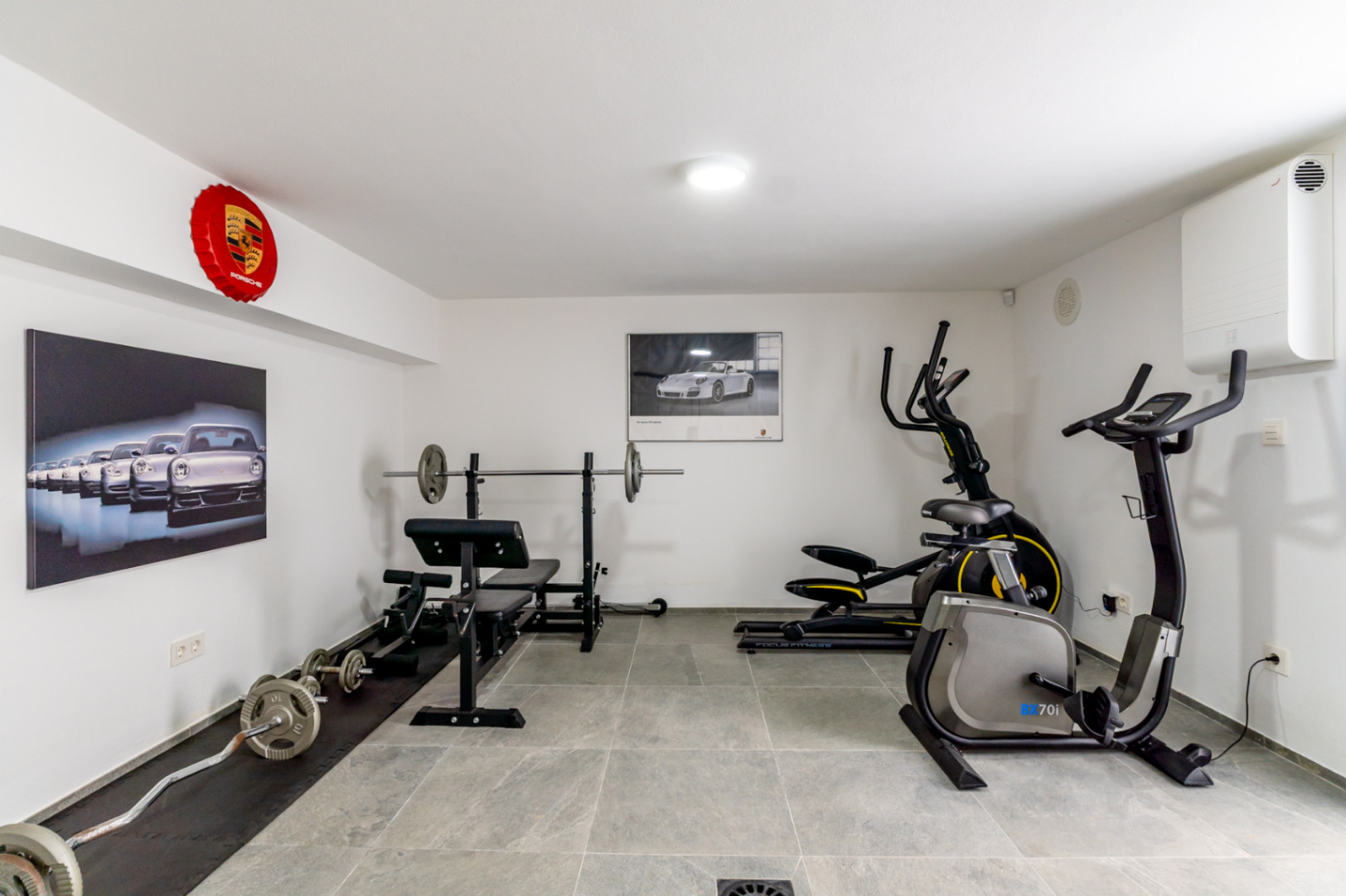 La salle de sport à domicile bien équipée comprend un banc de musculation, un vélo elliptique et un vélo stationnaire, sur un sol carrelé gris et décorée d'œuvres d'art Porsche.
