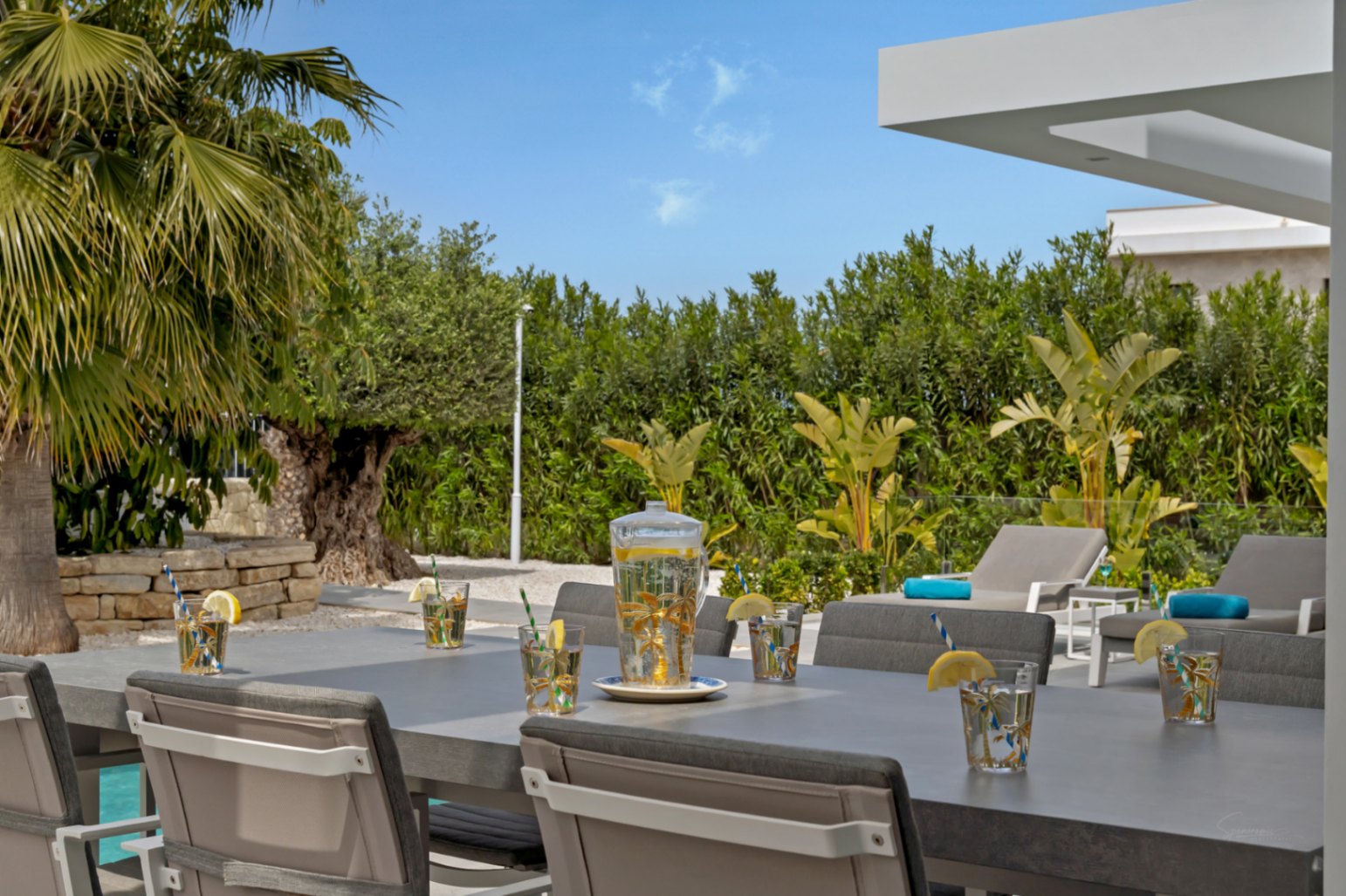L'espace repas extérieur dispose d'une table grise moderne avec des boissons rafraîchissantes, entouré d'un aménagement paysager méditerranéen luxuriant, de chaises longues et d'un aperçu de la piscine.