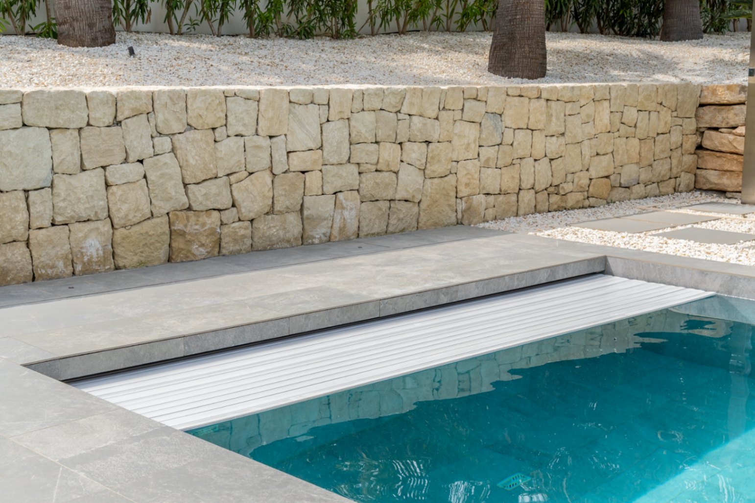 Piscine avec couverture à lames intégrée et margelle en carrelage gris moderne. Mur en pierre naturelle, gravier blanc et palmiers en arrière-plan.