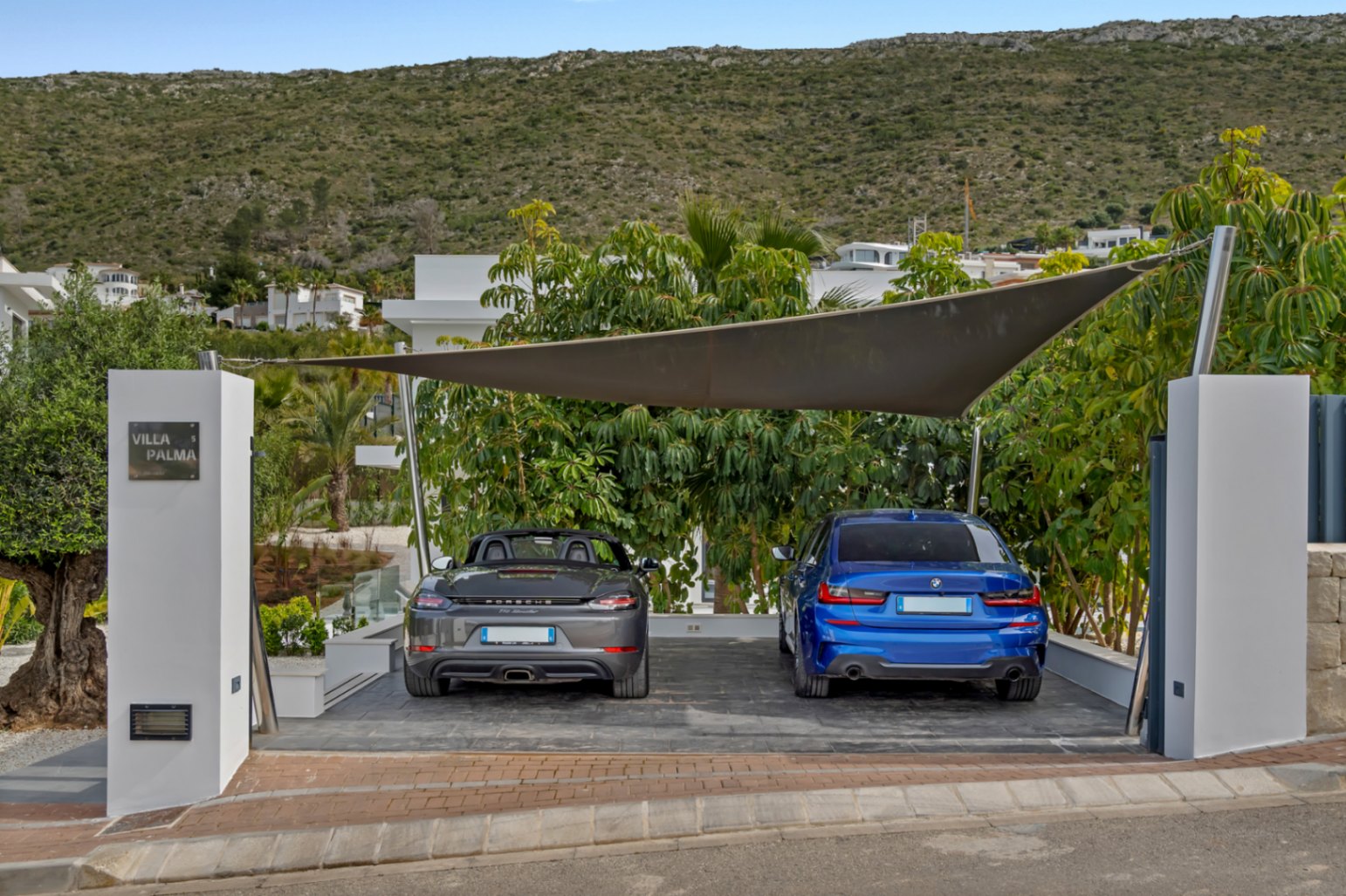 Place de stationnement extérieure sous voile d'ombrage moderne, abritant une Porsche grise et une BMW bleue. Le pilier d'entrée arbore l'enseigne 'Villa Palma', avec végétation méditerranéenne et colline en arrière-plan.