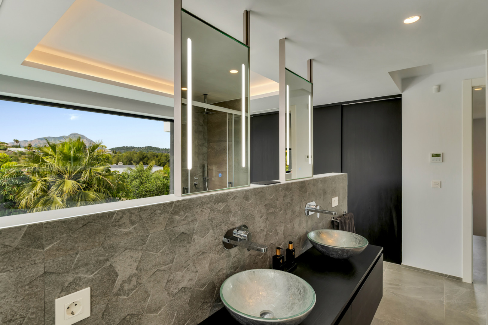 Salle de bain moderne avec double vasque en verre, crédence en carreaux hexagonaux gris texturés et miroirs suspendus éclairés. Grande fenêtre avec vue sur les montagnes verdoyantes.