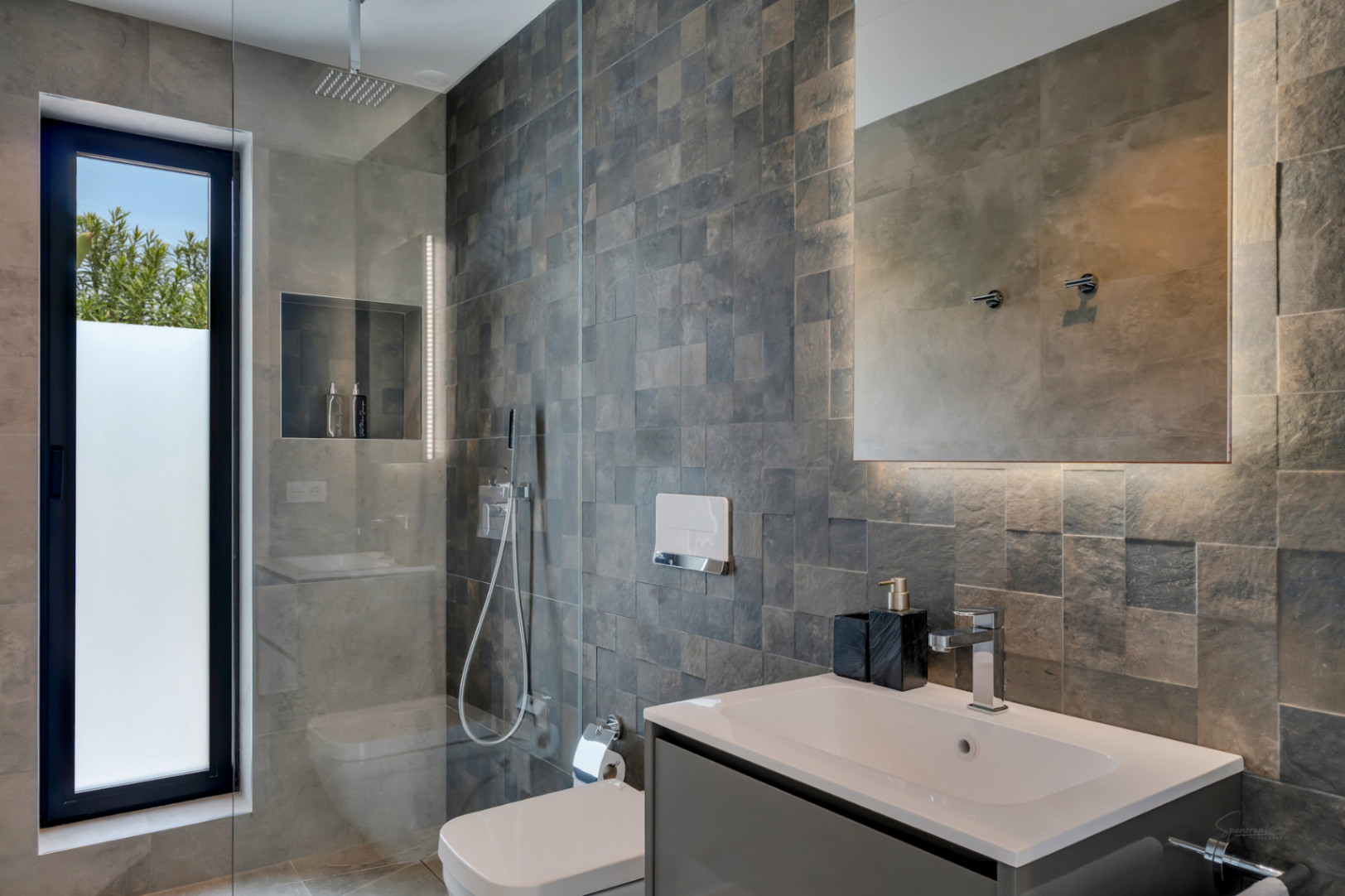 Salle de bain moderne avec douche à effet pluie, toilettes suspendues et meuble-lavabo gris. Les murs sont revêtus de carreaux texturés gris et une fenêtre verticale offre une vue sur la verdure.