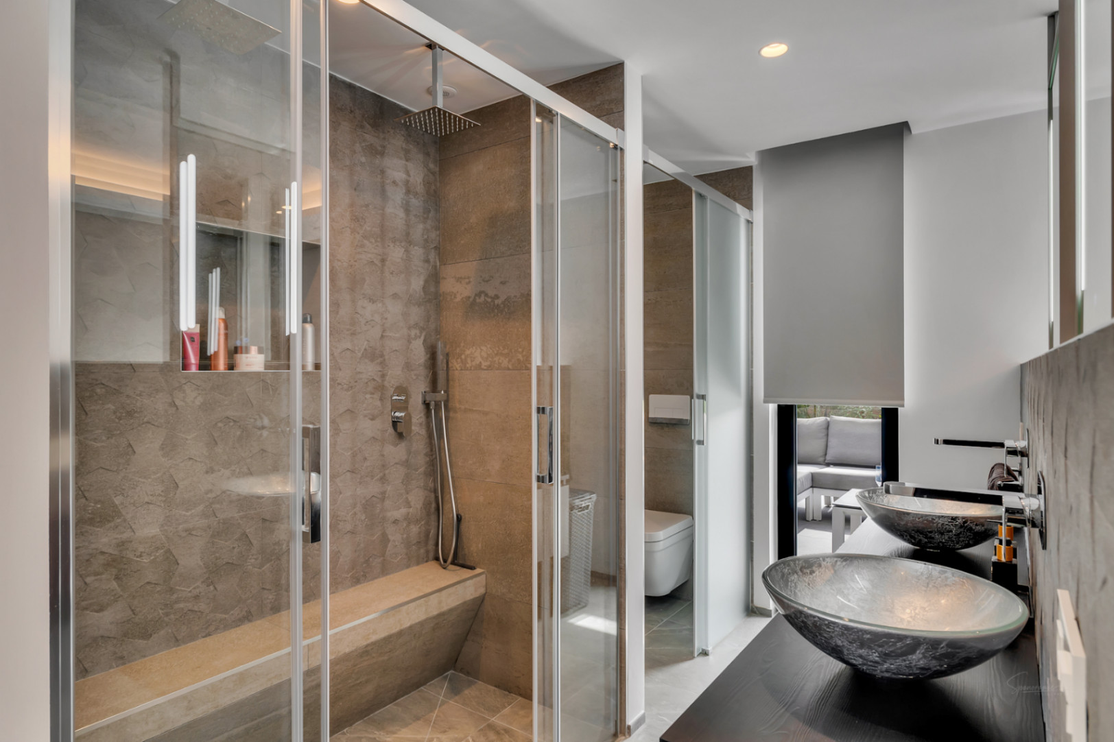 Salle de bain moderne avec une grande douche à l'italienne en pierre texturée, banc intégré et pommeau de pluie. Double vasque en verre sur meuble foncé, avec vue sur un salon extérieur.