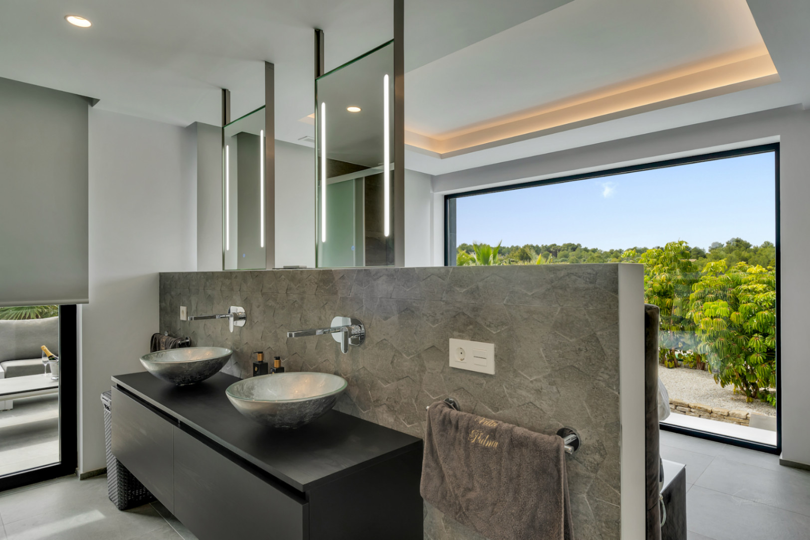 Salle de bain principale élégante avec double vasque design, mur en pierre texturée et miroirs éclairés par LED, offrant une vue dégagée sur la nature verdoyante.