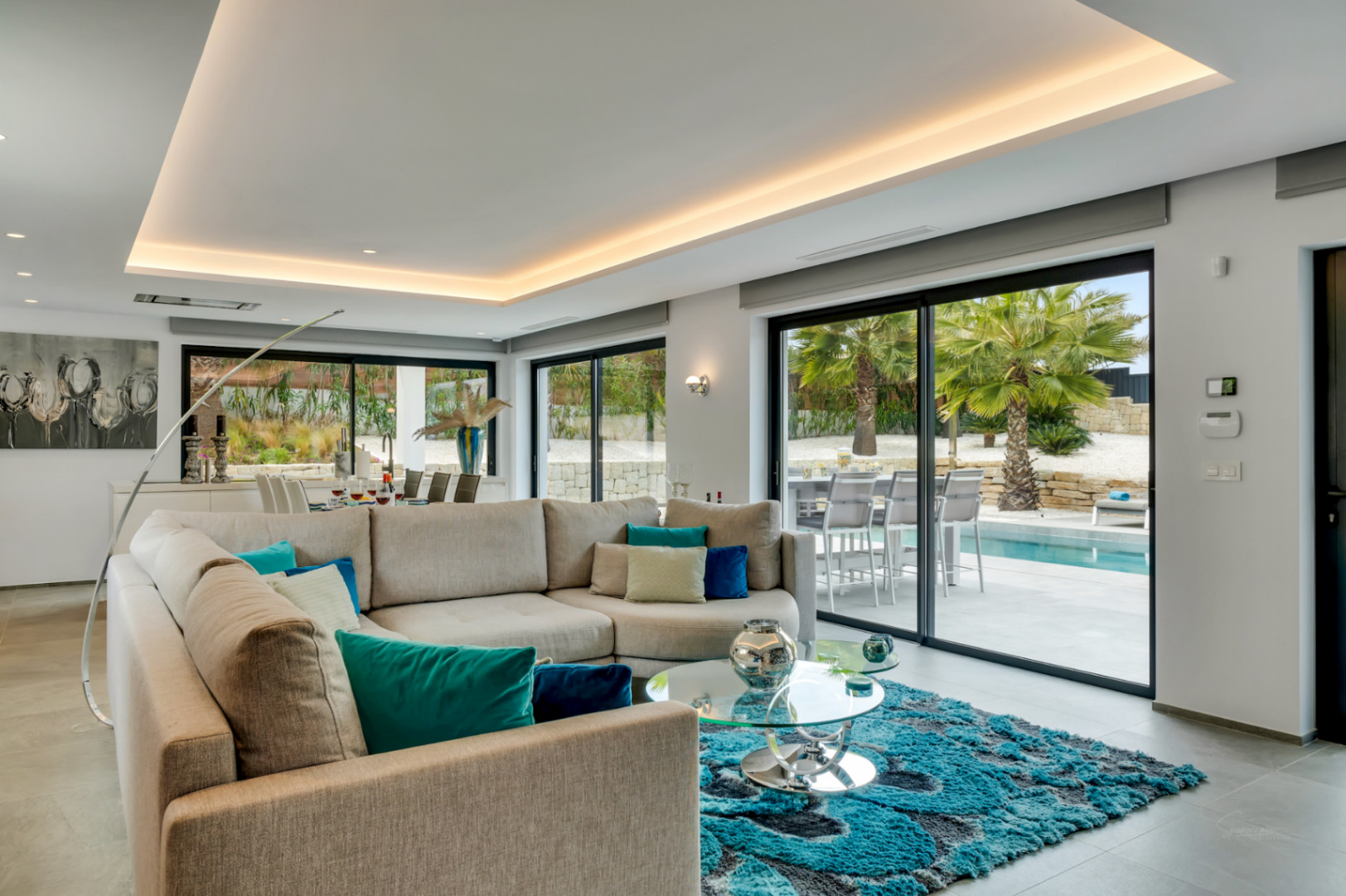 Salon moderne avec canapé en L, table basse en verre et tapis texturé. De grandes baies vitrées offrent un accès direct à la terrasse de la piscine et aux palmiers.