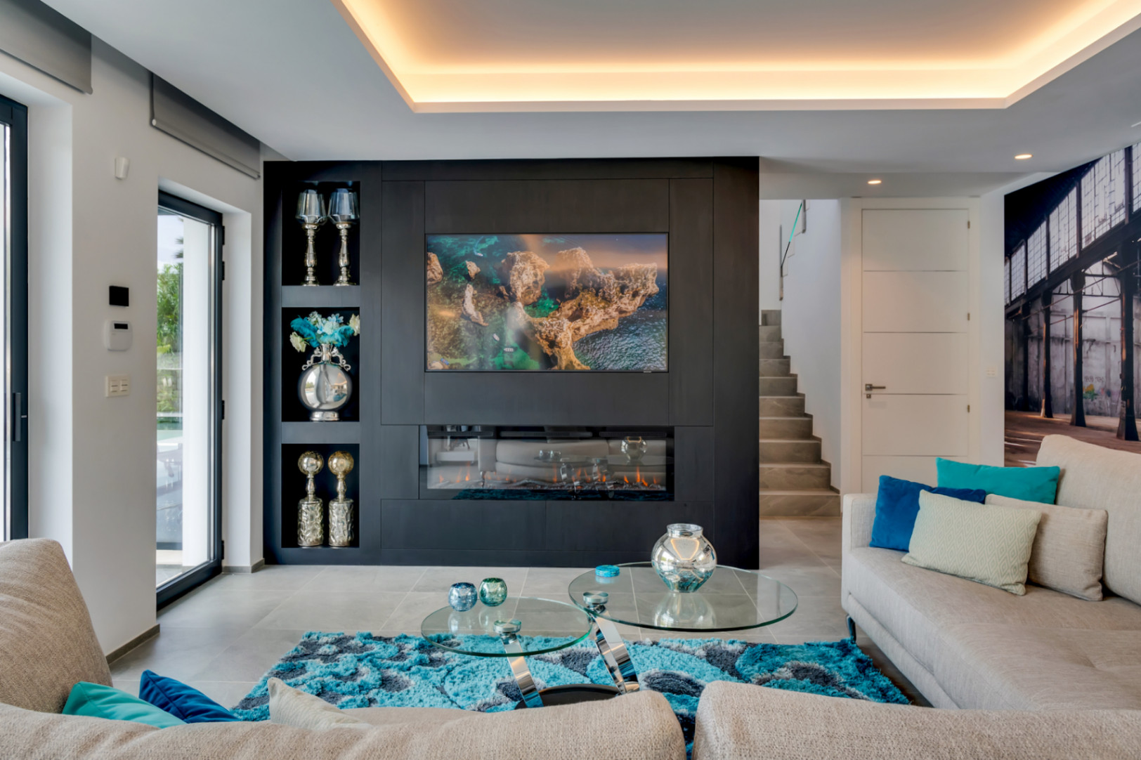 Salon moderne doté d'un mur d'accent sombre avec télévision et cheminée électrique intégrées, éclairage indirect et grandes portes vitrées vers l'extérieur.