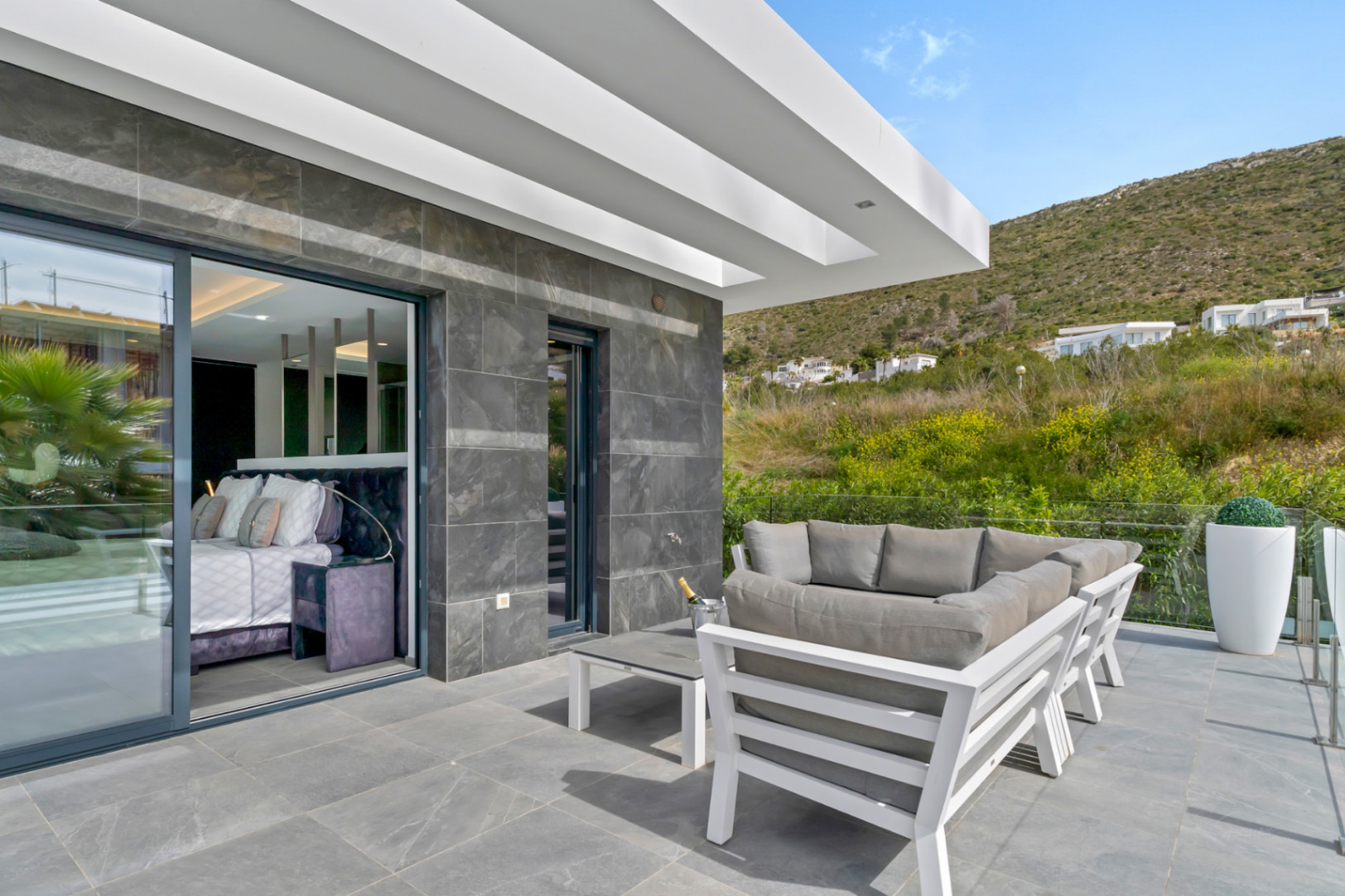 Terrasse extérieure carrelée de gris avec salon de jardin moderne, accès direct à une chambre et vue sur la colline verdoyante.