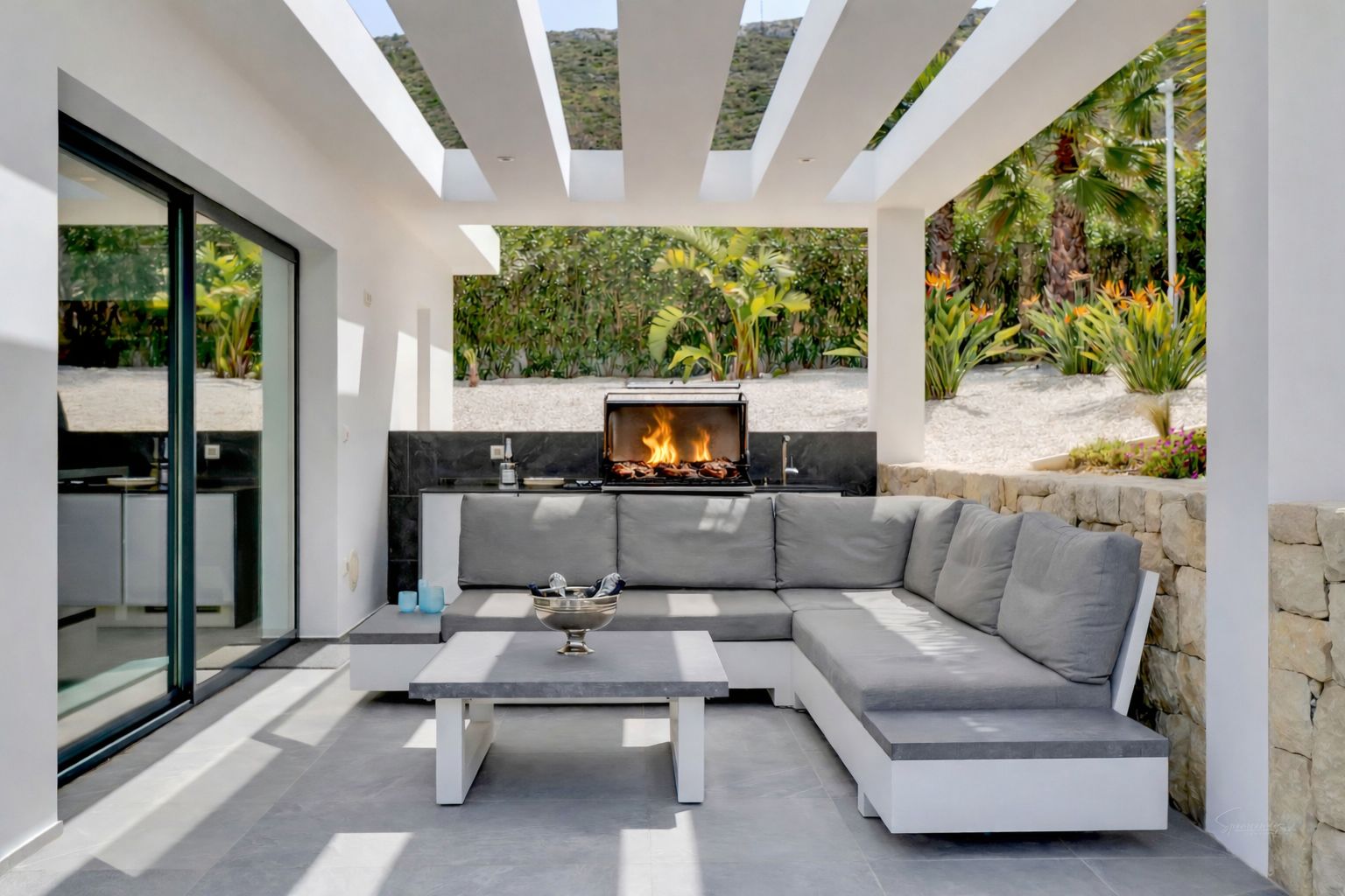 Terrasse extérieure couverte avec cuisine intégrée et barbecue allumé, complétée par un canapé lounge. Le plafond à lattes blanches projette des ombres sur le carrelage gris, face à un jardin méditerranéen luxuriant.