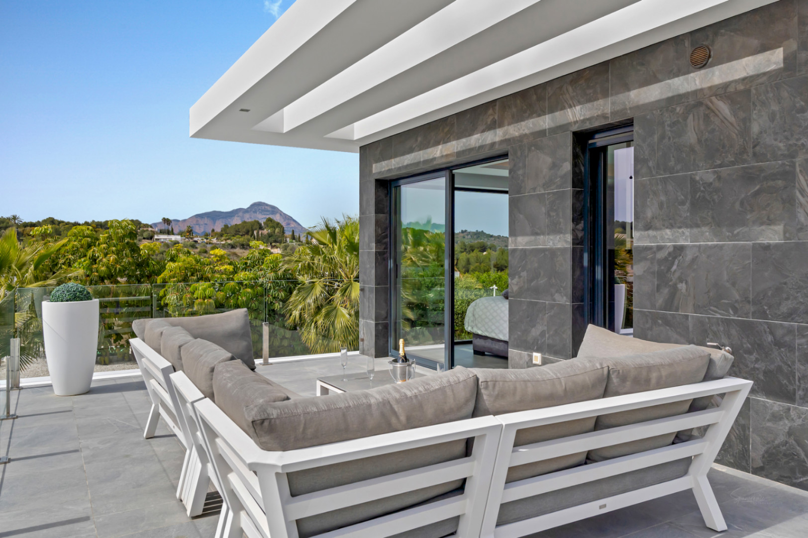 Terrasse extérieure moderne avec canapé gris en L, murs carrelés sombres et balustrade en verre. Vues sur les montagnes et la verdure, avec accès direct à une pièce intérieure.