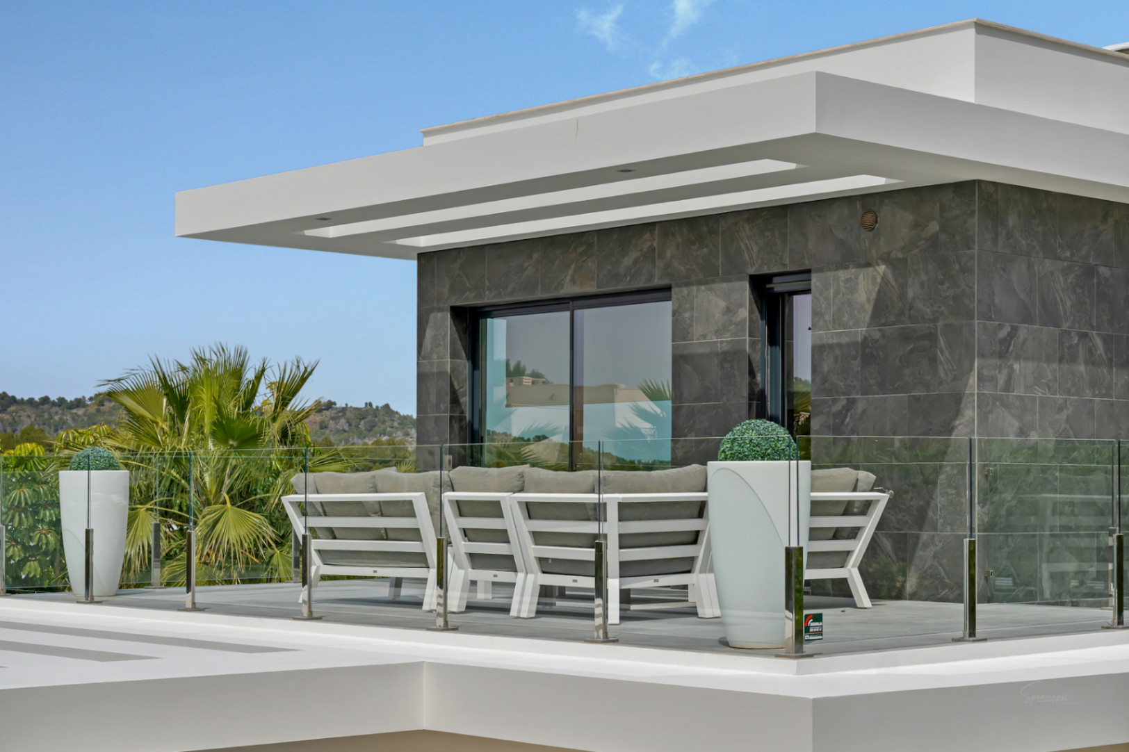 Terrasse extérieure moderne avec canapés blancs, garde-corps en verre et façade carrelée gris foncé. Vues sur la végétation luxuriante et les collines.