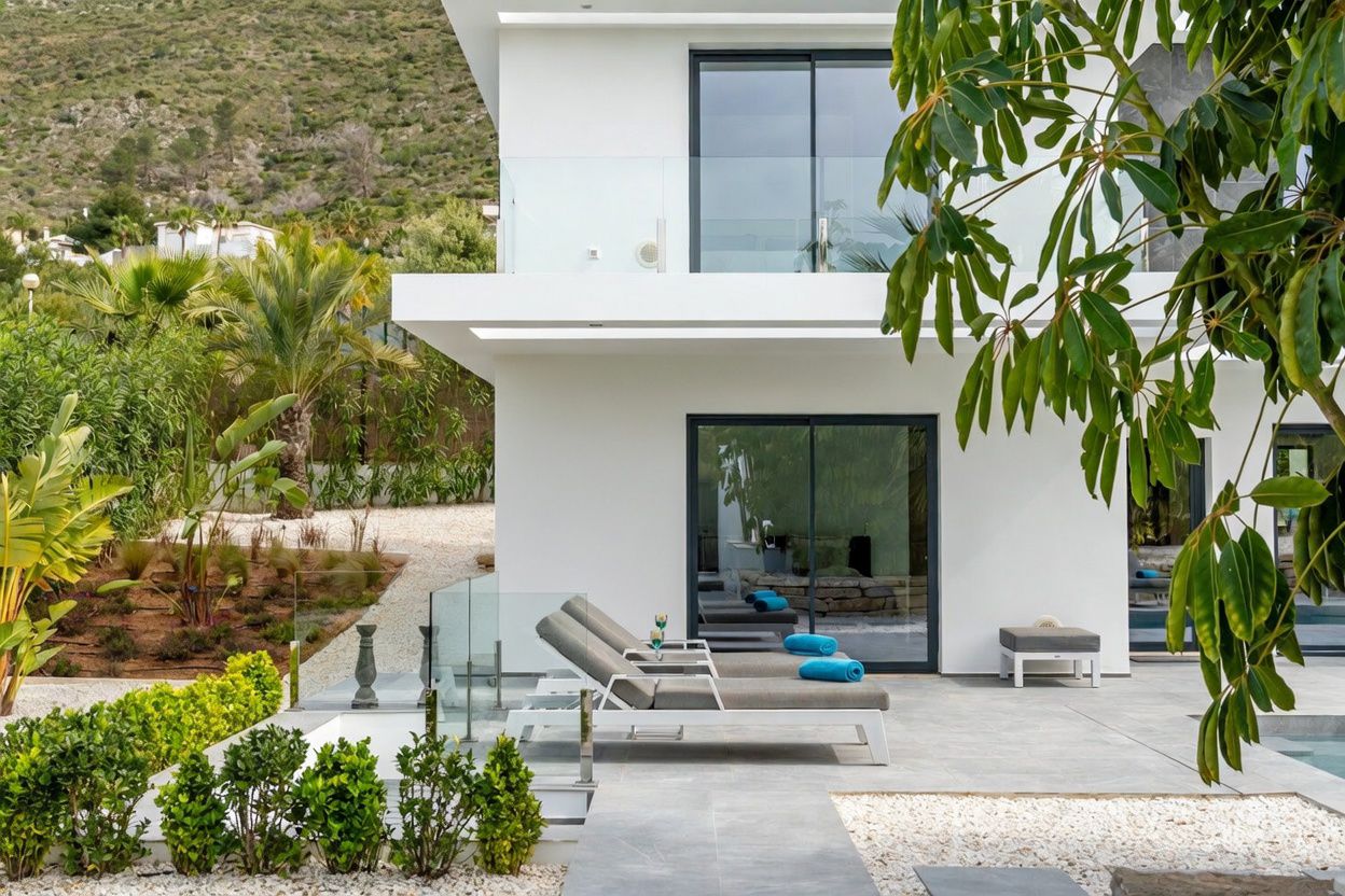 Terrasse moderne avec chaises longues grises sur carrelage grand format, délimitée par des balustrades en verre et un jardin paysager. Façade blanche avec portes coulissantes et fond montagneux.