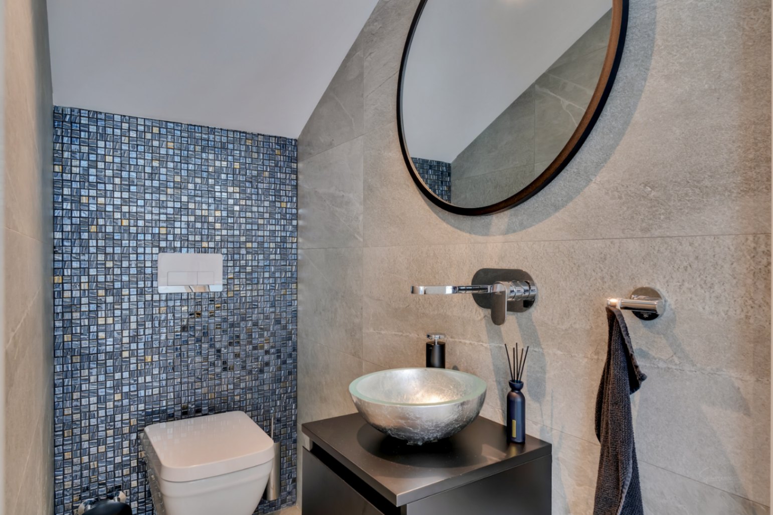 Toilettes d'invités modernes avec mur d'accent en mosaïque bleue et dorée, WC suspendu et lavabo en verre argenté sur meuble sombre.