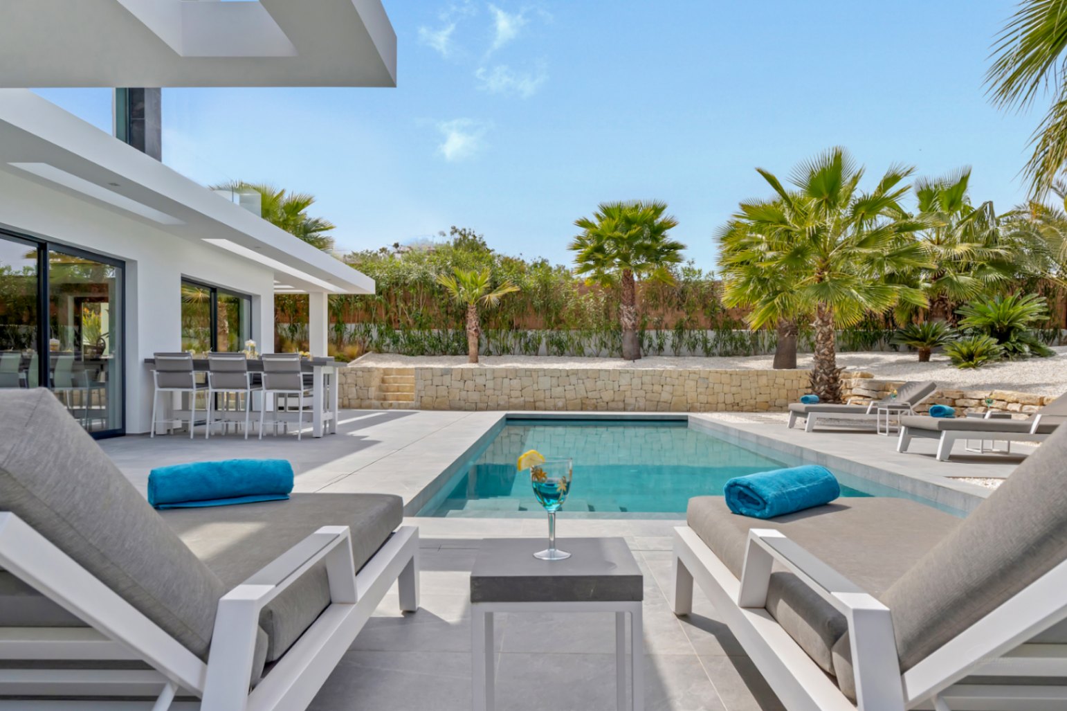 Vaste terrasse moderne avec piscine rectangulaire, chaises longues élégantes et bar extérieur intégré, reliée à la maison et aux palmiers.