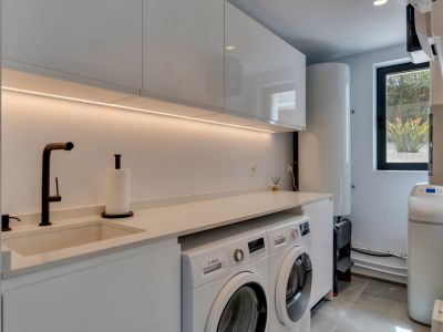 Buanderie moderne avec armoires blanches laquées, un évier intégré et des appareils Bosch. L'éclairage LED sous les meubles et une fenêtre donnent sur la végétation luxuriante.
