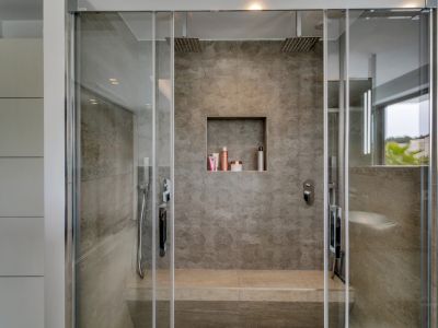 Cabine de douche moderne avec parois vitrées, carrelage gris effet pierre et deux pommes de douche pluie. Comprend un banc intégré, une niche encastrée et des rangements blancs. Vue partielle sur l'extérieur.