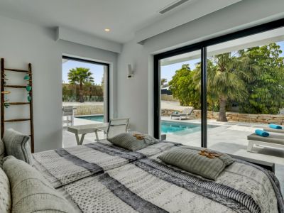 Chambre moderne avec accès direct à la piscine par de grandes baies vitrées coulissantes, révélant des transats, des palmiers et une végétation luxuriante.