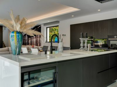 Cuisine ouverte moderne avec îlot en quartz blanc, armoires sombres et électroménager intégré, incluant une cave à vin, donnant sur un jardin verdoyant.