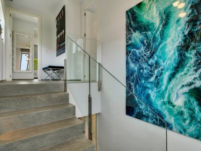 Escalier en pierre avec balustrade en verre, mis en valeur par une grande œuvre d'art abstraite bleu-vert. Un couloir avec miroir décoratif et banc est visible à l'étage.