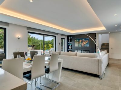 Espace de vie et salle à manger ouvert moderne avec table blanche élégante, canapé gris clair et mur média intégrant une cheminée électrique. Larges baies vitrées donnent sur le jardin.