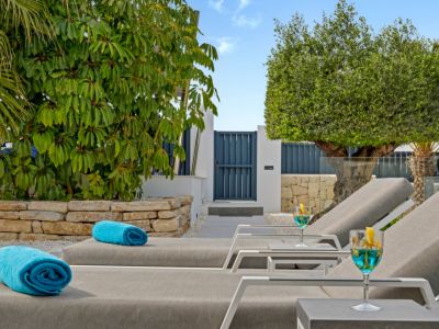 Espace détente extérieur avec deux chaises longues modernes, serviettes bleues et cocktails. Un chemin mène à un portail bleu, encadré de verdure luxuriante et d'un mur de pierre.
