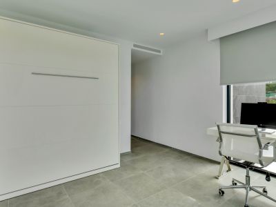 Espace multifonctionnel avec lit escamotable intégré, bureau moderne devant une fenêtre avec store et carrelage gris au sol.