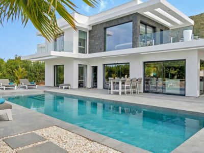 Extérieur de villa moderne avec piscine rectangulaire et vaste terrasse carrelée. De grandes baies vitrées relient l'intérieur, avec un espace repas extérieur et des vues sur la montagne.
