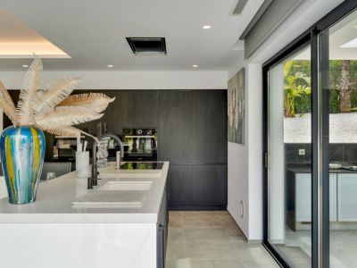 Îlot de cuisine moderne avec plan de travail blanc et éviers intégrés. Armoires sombres minimalistes avec appareils encastrés, et portes vitrées coulissantes vers la cuisine extérieure et le jardin.