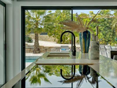 Îlot de cuisine moderne avec surface réfléchissante et robinet noir épuré. De grandes baies vitrées offrent des vues sur les palmiers, les murs en pierre et la piscine, connectant intérieur et extérieur.