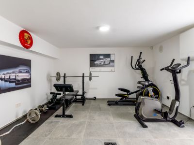 La salle de sport à domicile bien équipée comprend un banc de musculation, un vélo elliptique et un vélo stationnaire, sur un sol carrelé gris et décorée d'œuvres d'art Porsche.