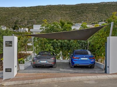 Place de stationnement extérieure sous voile d'ombrage moderne, abritant une Porsche grise et une BMW bleue. Le pilier d'entrée arbore l'enseigne 'Villa Palma', avec végétation méditerranéenne et colline en arrière-plan.
